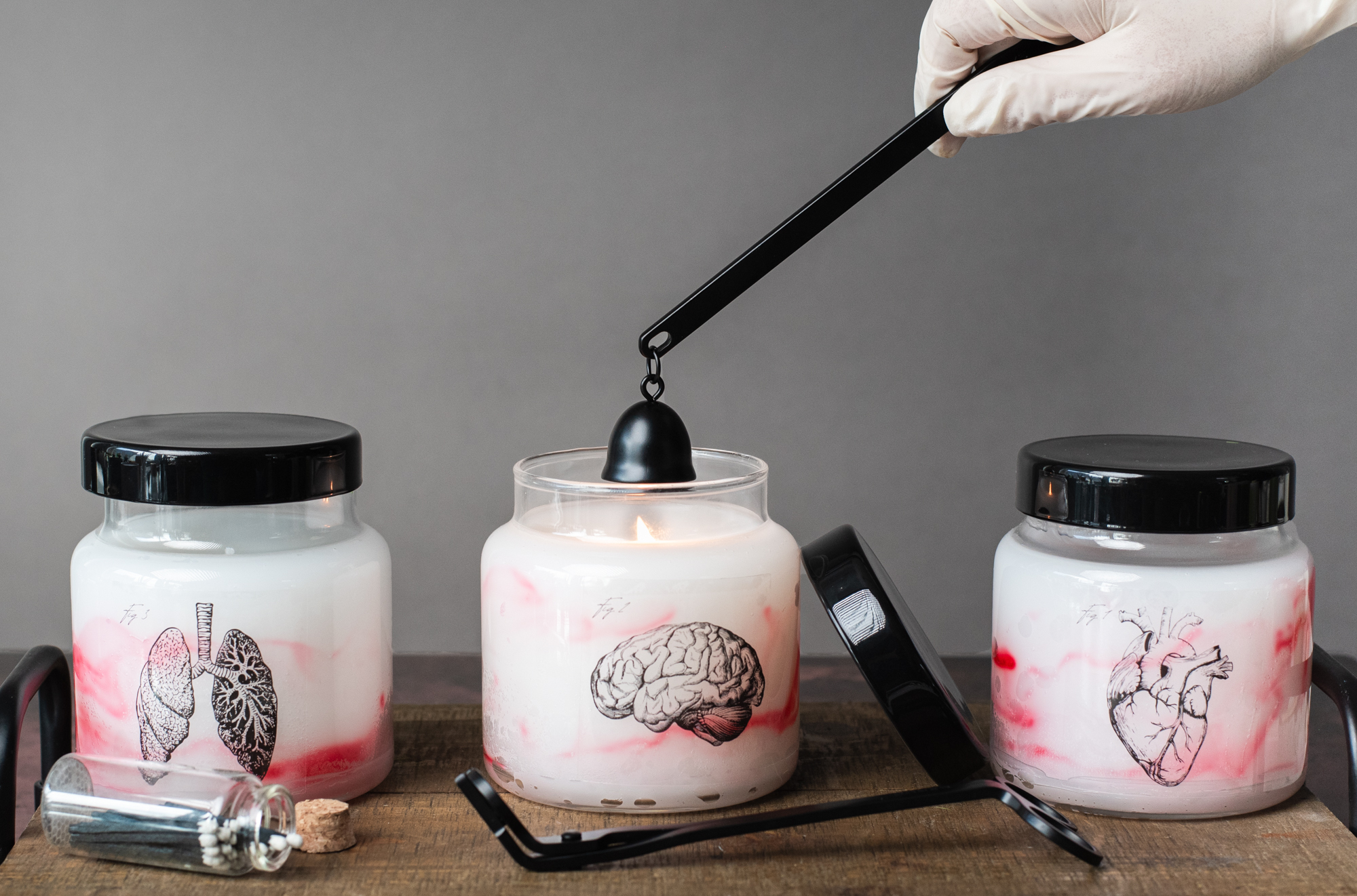 Three candles in a row made in 16 oz glass apothecary jars with black glass lids. The candles are dyed red with a marbled technique. Images of lungs, a brain, and a heart are on the outside of the candles. A gloved hand reaches into the frame with a black candle snuffer to extinguish the center candle.